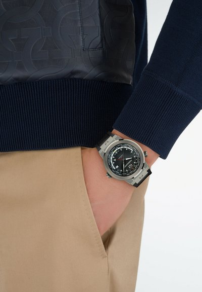 Pull en tricot bleu avec des poignets côtelés, associé à un pantalon beige. Une grande montre-bracelet en argent et noir présente un cadran rond et plusieurs cadrans.