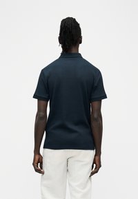 Homme de dos portant un polo côtelé à manches courtes bleu marine et un pantalon blanc, sur un fond clair uni.