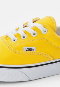 Gul kanvas sneakers med vita snören, svart gummisula och en liten vit Vans-logotagg. Har en slät yta och klassisk lågtoppsdesign.