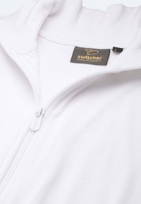 Weiße Zip-Hoodie aus weichem Stoff mit hohem Kragen und einem Etikett mit "Switcher"-Logo und Nachhaltigkeitsbotschaft. Reißverschluss ist grau.