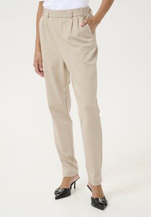Pantalons beige ajustés avec des plis, taille haute et poches latérales. Portés avec des chaussures à talons noires ornées d'un nœud décoratif.