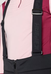 Czarne regulowane szelki o teksturowanej powierzchni, przypięte do dopasowanej bluzki w jasnym różu i ciemnym burgundzie; zamek błyskawiczny z przodu w talii.