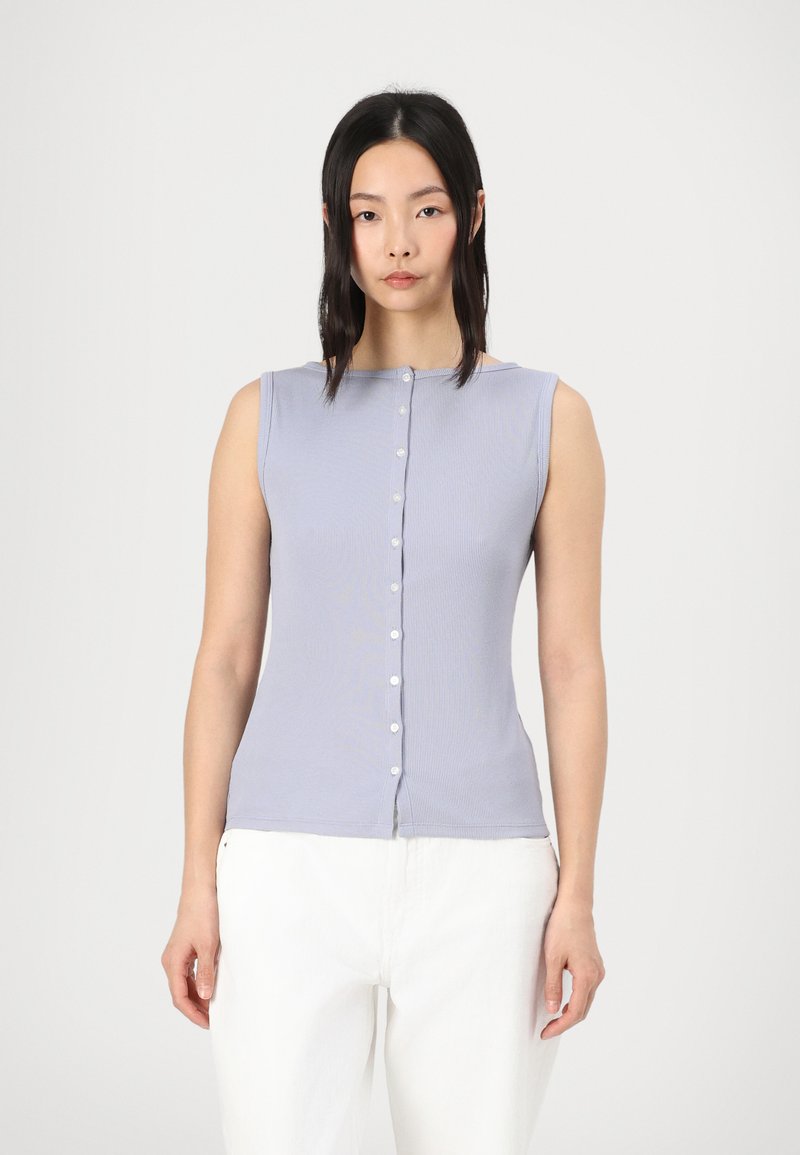 Top sans manches de couleur violet clair avec une texture côtelée et une fermeture à boutons sur le devant. Associé à un pantalon blanc ample.