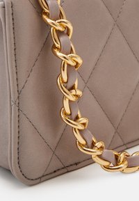 Borsa a tracolla trapuntata color taupe con catena dorata intrecciata attraverso la tracolla in pelle coordinata su sfondo bianco.