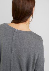 Harmaa neulepaita, jossa on pehmeä rakenne, leveä kaula-aukko ja nappikiinnitys selkäpuolella. Yksinkertainen muotoilu ilman kuvioita.