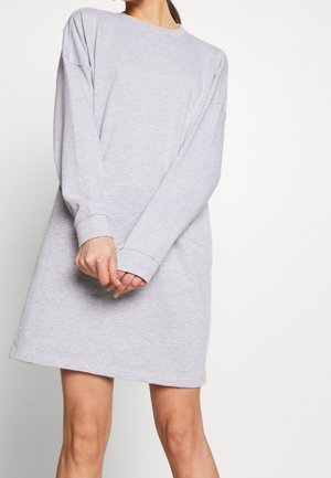 Frau trägt ein hellgraues Sweatshirtkleid mit langen Ärmeln, steht mit vor dem Körper verschränkten Händen vor einem schlichten weißen Hintergrund.