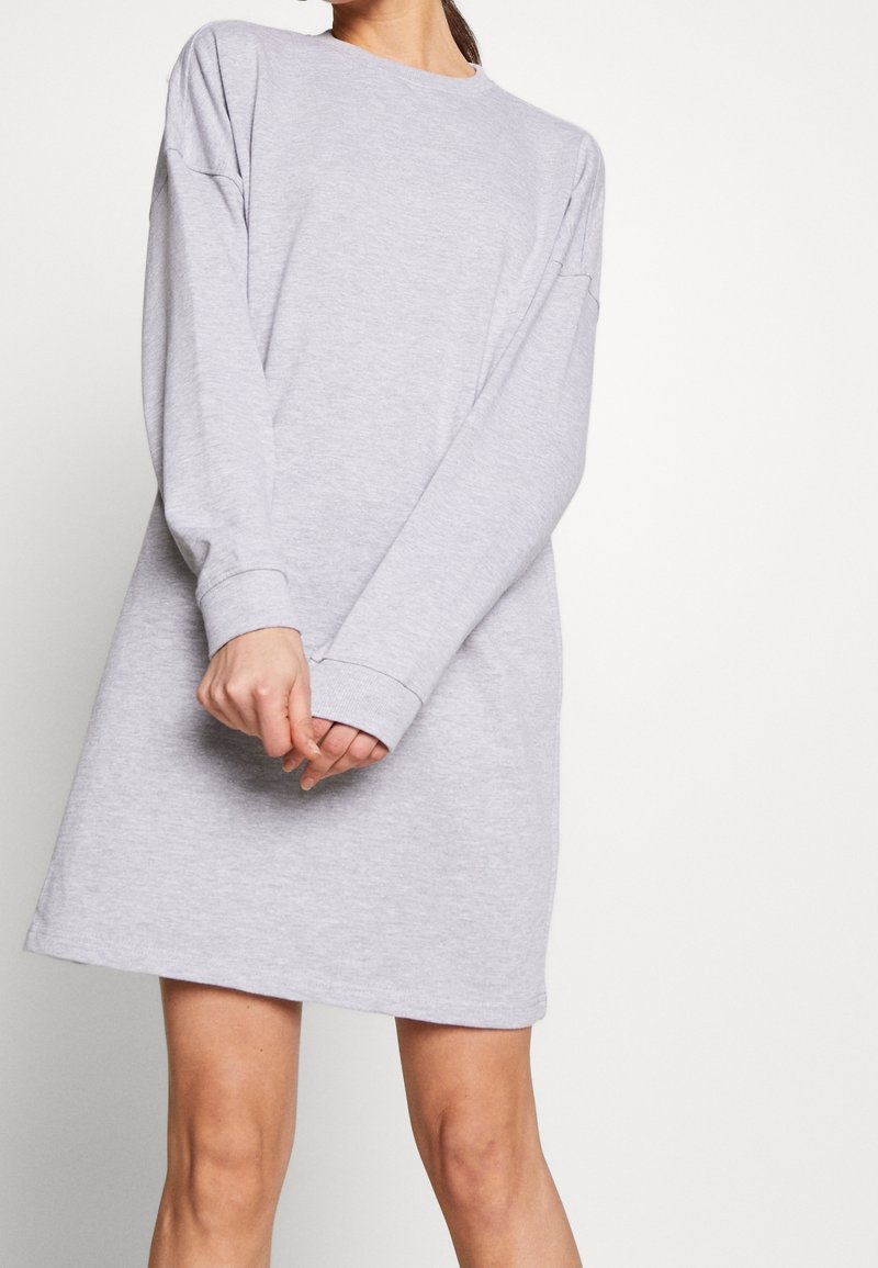 Femme portant une robe sweat-shirt à manches longues gris clair, debout les mains jointes devant un fond blanc uni.