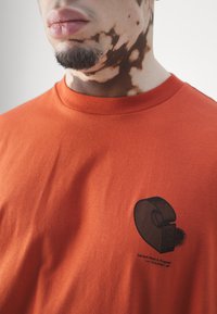Camiseta de algodón naranja con cuello redondo, que presenta un diseño gráfico negro de cinta y texto en la parte inferior izquierda. Textura suave.