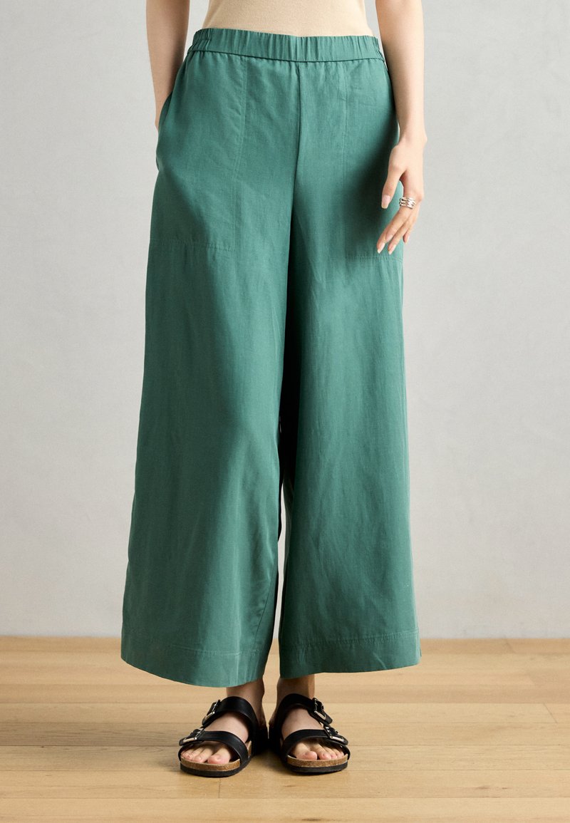 Pantalons larges verts avec une ceinture, présentant une texture lisse et des poches latérales. Portés avec des sandales noires ornées de boucles.