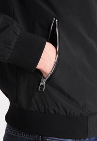 Černá bomber bunda s kapsou na zip, elastickými manžetami a hladkou texturou. Design zahrnuje přiléhavý pás a minimální detaily.