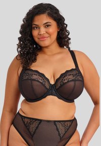 Knalalhaarige vrouw in een zwarte kanten en netten beha en bijpassende high-waisted slipjes, lachend tegen een effen grijze achtergrond.