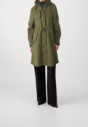 Femme portant un manteau à capuche vert olive avec poches devant, taille avec cordon, pantalon large noir et chaussures noires à bout pointu.