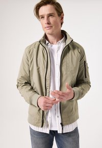 Chaqueta bomber en color caqui claro con cuello alto, bolsillos laterales y cierre de cremallera negro; llevada sobre una camisa blanca de botones con rayas azules.