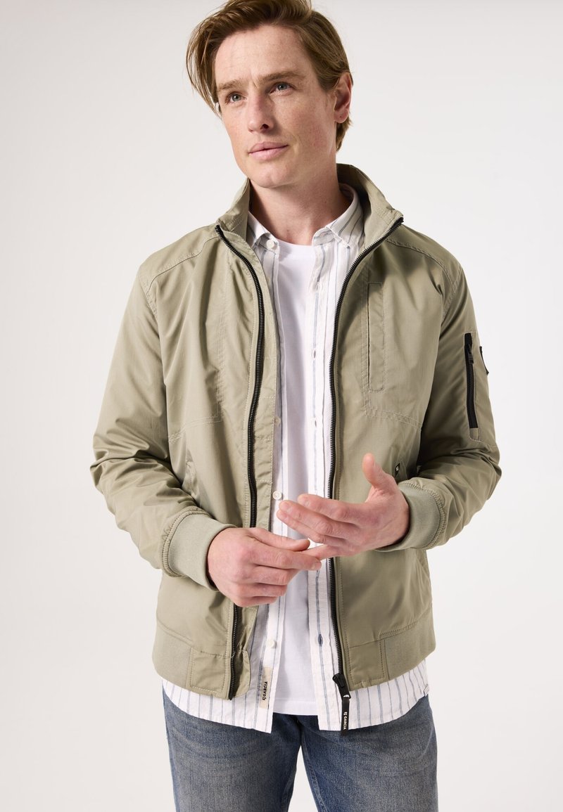 Chaqueta bomber en color caqui claro con cuello alto, bolsillos laterales y cierre de cremallera negro; llevada sobre una camisa blanca de botones con rayas azules.