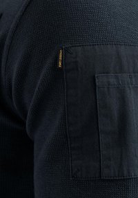 Schwarzes, strukturiertes Stoffhemd mit einer aufgesetzten Tasche am Ärmel und einem kleinen gelben Etikett mit der Aufschrift "PHE LEGEND".