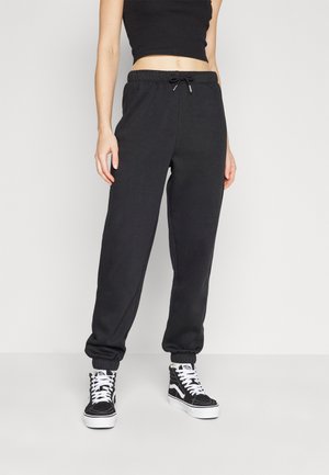 Femme portant un débardeur court noir, un pantalon de jogging à taille élastique noir et des baskets montantes noires avec des lacets blancs, debout contre un mur blanc.