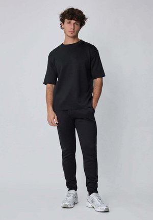 Jeune homme aux cheveux bouclés portant un t-shirt noir oversize, un pantalon slim noir et des baskets blanches, debout devant un fond uni clair.