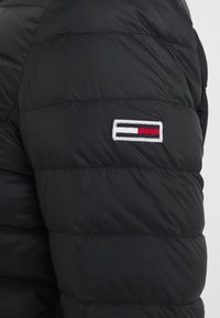 Doudoune noire à motif matelassé. Présente un logo brodé avec des rayures rouge, blanc et bleu sur la partie supérieure du bras. Texture lisse.