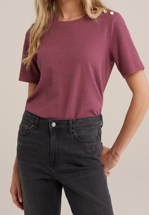 Kvinde iført en kortærmet lilla top med guldknapper på skulderen, stoppet ned i højtaljede sorte jeans, hånd i lommen.