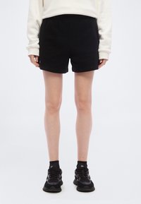 Άτομο που φορά μαύρο αθλητικό σορτς, μαύρα αθλητικά παπούτσια με κάλτσες και ανοιχτόχρωμο μπλουζάκι με μακριά μανίκια, στέκ εται πάνω σε απλό λευκό φόντο.