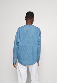 Camisa de mezclilla de color azul claro con corte relajado, mangas largas y dobladillo curvado. Presenta un cuello sencillo y puños abotonados. Textura suave.