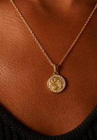 Gouden ketting met een ronde hanger, voorzien van een ingewikkeld ontwerp en een rand van heldere kristallen, rustend op een gestructureerd huidoppervlak.