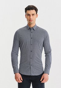 Mężczyzna stojący przodem, ubrany w dopasowaną koszulę z długim rękawem, z guzikami, w mały geometryczny wzór oraz ciemne spodnie.