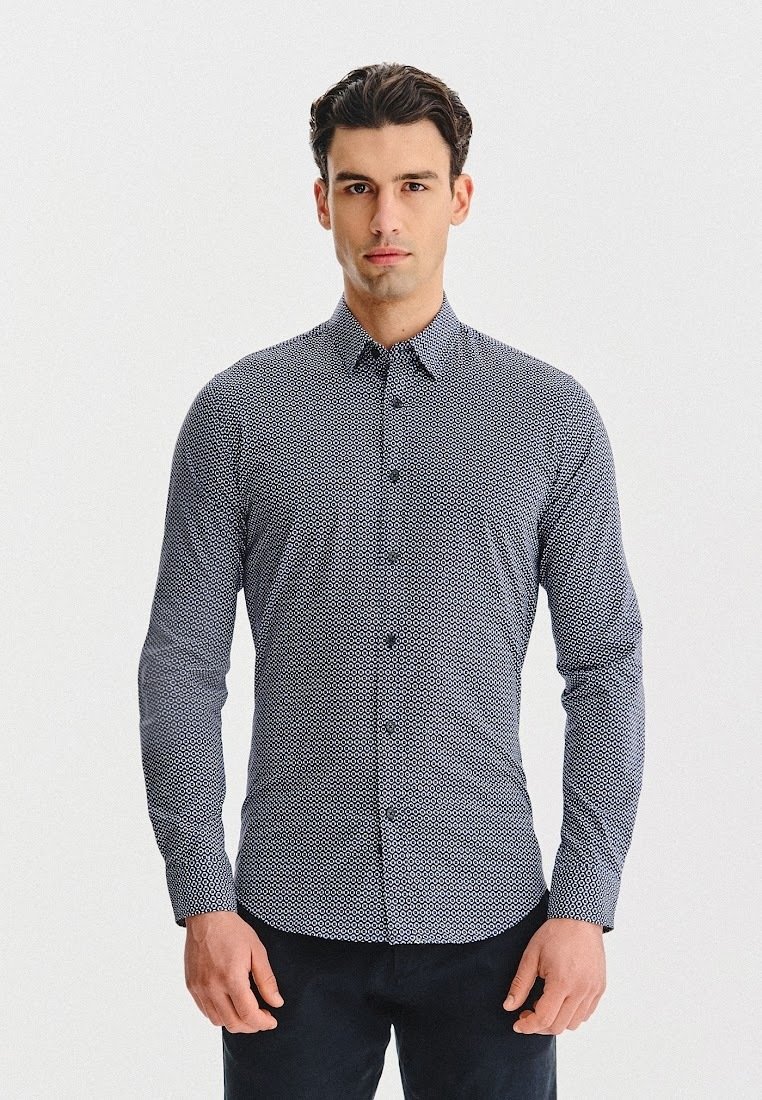 Mężczyzna stojący przodem, ubrany w dopasowaną koszulę z długim rękawem, z guzikami, w mały geometryczny wzór oraz ciemne spodnie.