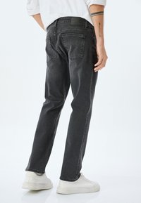 Vaqueros de denim negro con corte recto, bolsillos traseros visibles y una sutil textura desgastada, combinados con zapatillas blancas.