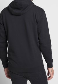 Felpa nera con cappuccio realizzata in tessuto morbido, dotata di cappuccio con cordoncino, polsini e orlo a coste. Design semplice, senza grafiche o motivi visibili.