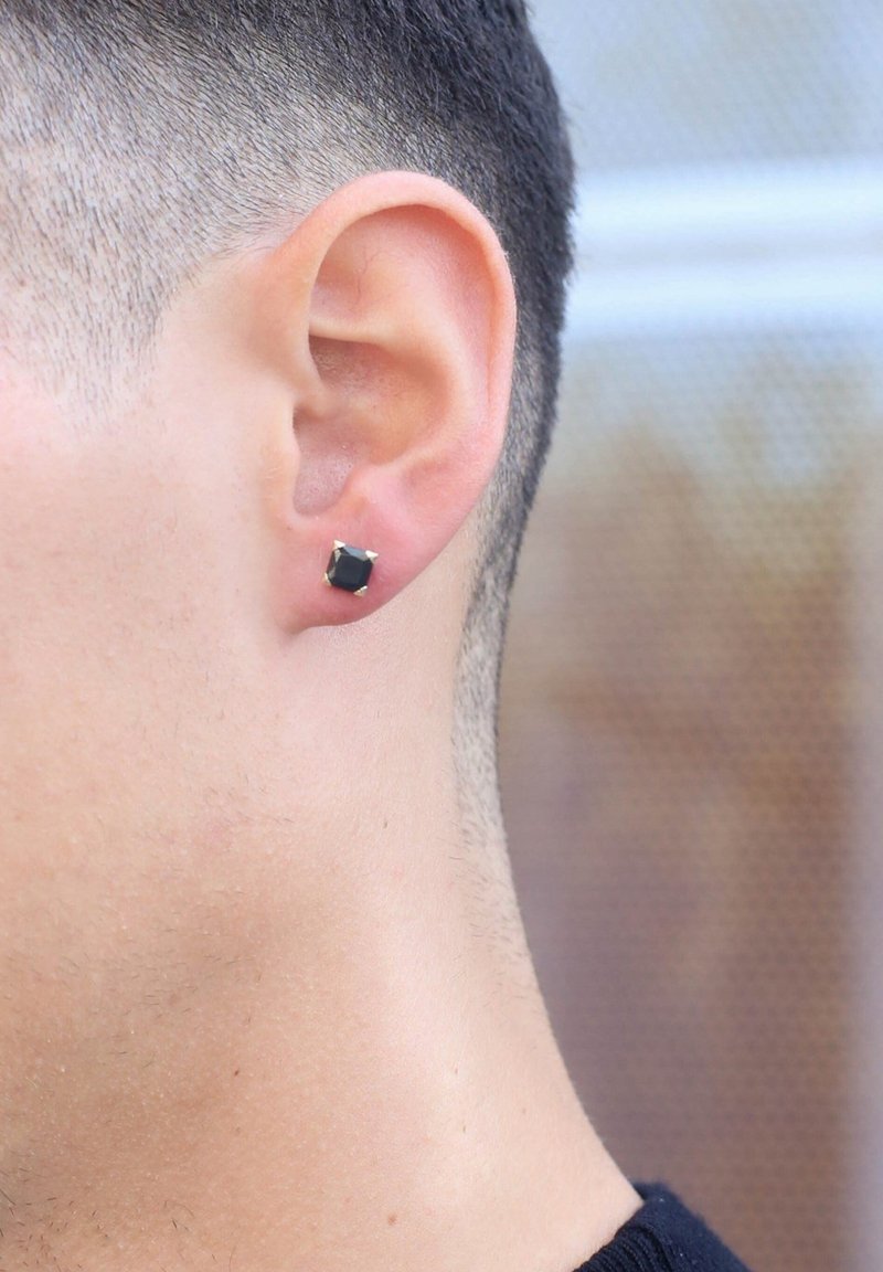 Boucle d'oreille noire carrée avec des accents dorés, attachée à l'oreille d'une personne. Gros plan sur la forme et la couleur de la boucle.