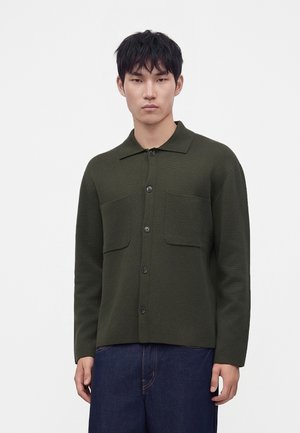 Homme portant une chemise à boutons vert foncé avec deux poches poitrine et un pantalon bleu foncé, debout devant un fond clair uni.