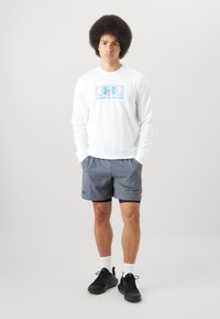 Camisa blanca de manga larga con el logo azul de Under Armour, combinada con pantalones cortos atléticos grises. Zapatillas negras, calcetines blancos. Diseño deportivo simple.