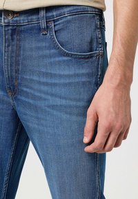 Jeans in denim blu con una vestibilità slim, cuciture visibili, tasca frontale e chiusura con bottone. Tessuto liscio con una finitura leggermente sbiadita.