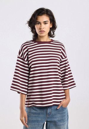 Jong persoon met kort golvend haar, gekleed in een losse bordeauxrode en witte gestreepte blouse en blauwe jeans, staand met één hand in de zak.