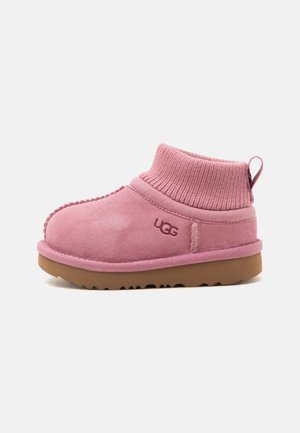 Chausson UGG en daim rose à la cheville avec bord-côte tricoté et semelle en caoutchouc marron, vue de côté montrant le logo UGG sur le côté extérieur.