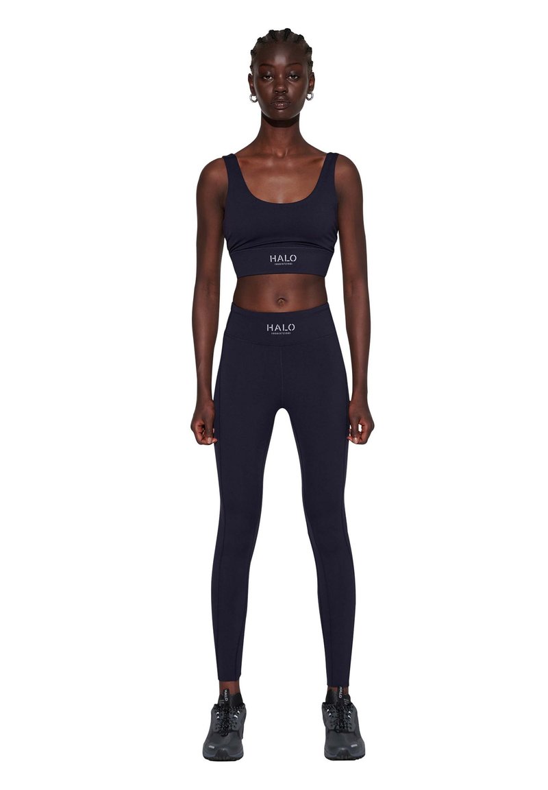 Marineblauwe sport crop top en leggings gemaakt van rekbare stof, met het "HALO" logo op de tailleband, gecombineerd met zwarte sportschoenen.