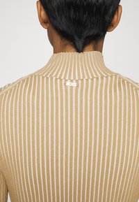 Maglione beige a coste con strisce verticali bianche, colletto alto e logo 'BOSS' sulla schiena. Texture morbida e design aderente.