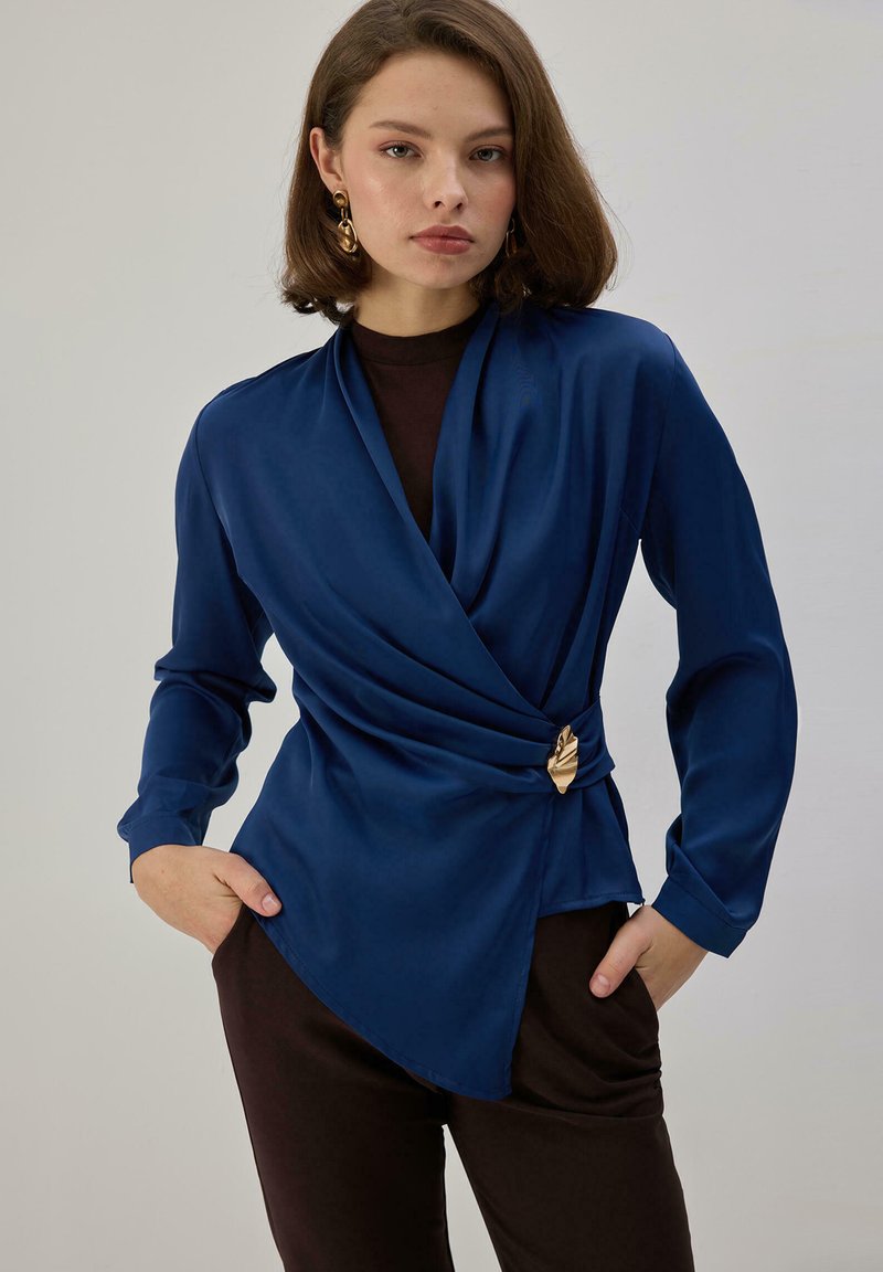 Blusa cruzada azul marino en tejido sedoso, con diseño drapeado, mangas largas y un detalle de hoja dorada en la cintura.