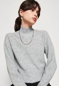 Suéter cinza tricotado com gola alta, textura canelada e ombros caídos. Os acessórios incluem um colar de corrente prateada e brincos pendentes.