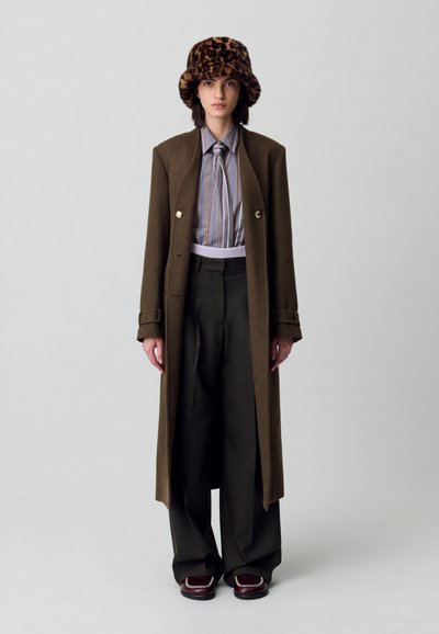 Cappotto di lana marrone con bottoni dorati, camicia a righe viola, pantaloni verdi larghi e un cappello in pelliccia sintetica a stampa leopardata. Scarpe borgogna con accenti bianchi.