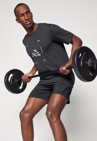 Camiseta atlética negra con un diseño gráfico, combinada con pantalones cortos negros. Persona levantando una barra con discos de pesas negros. Fondo sólido.