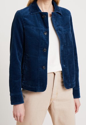 Kvinde iført en blå fløjlsjakke med knapper over en hvid tekstureret top og beige bukser, vist fra hals til hofter.