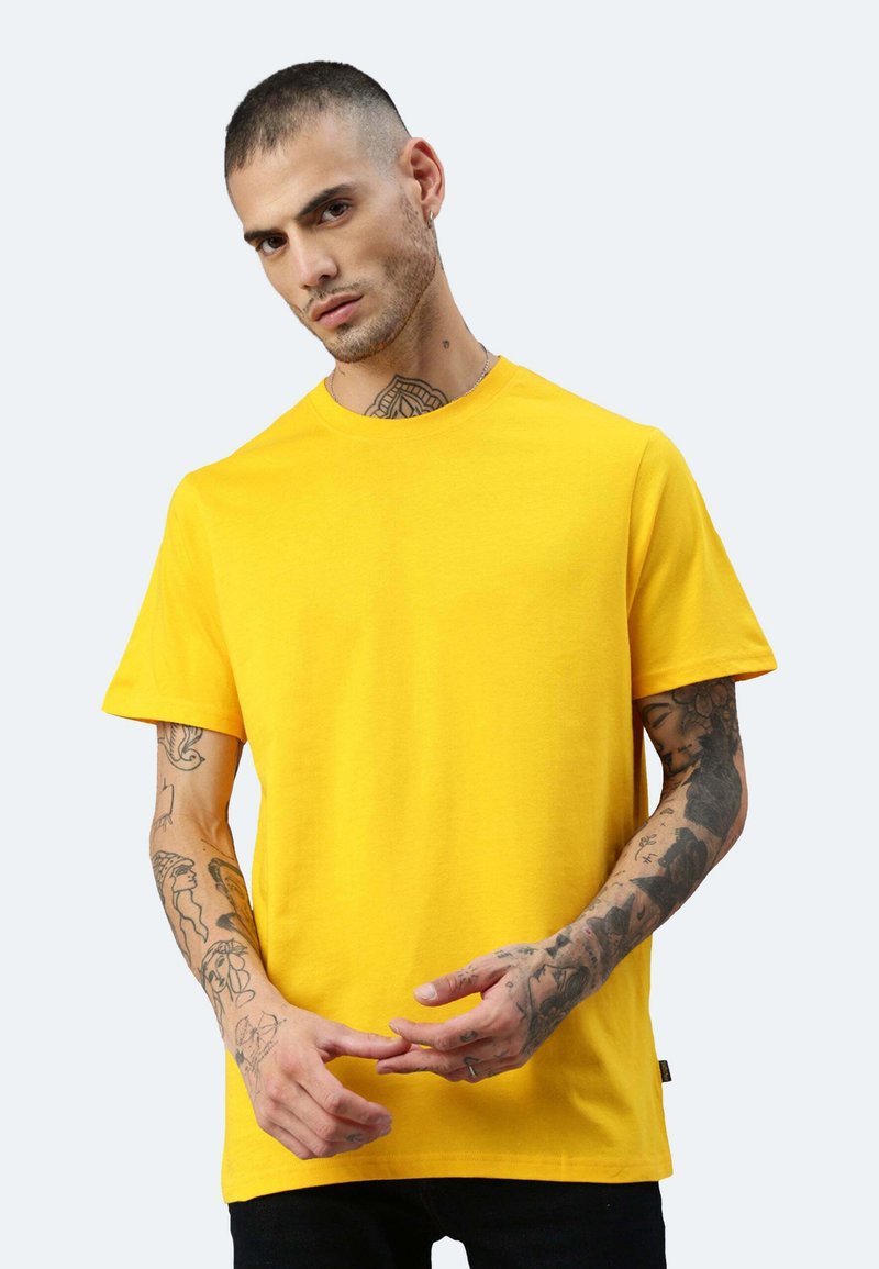 Gelbes Baumwoll-T-Shirt mit kurzen Ärmeln, Rundhalsausschnitt und schlichtem Design. Das Modell posiert mit sichtbaren Tätowierungen an den Armen.