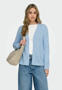 Woman with long blonde hair wearing a light blue knit cardigan over white top and wide-leg blue jeans, holding a beige shoulder bag.