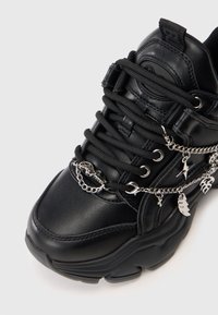 Zwarte vetersneaker met een robuuste zool, voorzien van zilveren kettingaccenten en decoratieve charms. Gemaakt van soepel leer met textuurdetails.