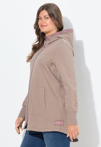 Giovane donna che indossa un felpa con zip color taupe con simboli di infinito rosa sulla cappuccio e sulla tasca, abbinata a jeans blu, in piedi di lato.