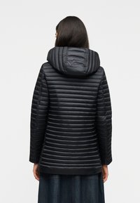 Γυναίκα φοράει μια μαύρη μπουφάν με κουκούλα και ραφή, φαίνεται από πίσω, συνδυασμένη με μια σκουρόχρωμη φούστα, σε απλό ανοιχτό φόντο.