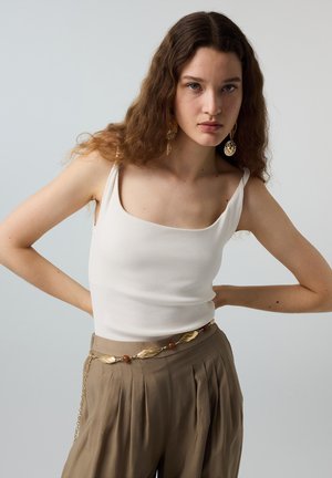 Jonge vrouw met lang krullend haar draagt een witte mouwloze top, geplooide bruine broek, gouden bladeren kettingriem en grote gouden oorbellen, staand met handen in de zij.