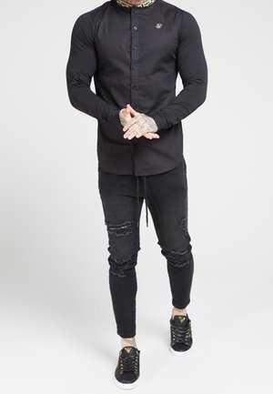 Mand iført sort skjorte med knapper og mønstret krave, sorte revnede jeans og sorte sneakers med gyldne accenter, der står mod hvid baggrund.